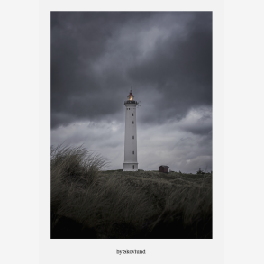 Lyngvig Fyr st�r oplyst mod en m�rk, dramatisk himmel med bl�sende marehalm i forgrunden og et roligt lys i t�rnets top.