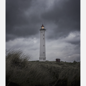 Lyngvig Fyr st�r oplyst mod en m�rk, dramatisk himmel med bl�sende marehalm i forgrunden og et roligt lys i t�rnets top.