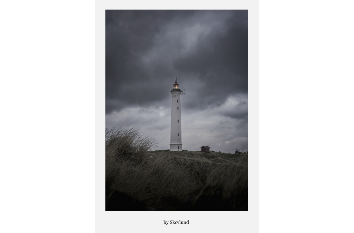 Lyngvig Fyr st�r oplyst mod en m�rk, dramatisk himmel med bl�sende marehalm i forgrunden og et roligt lys i t�rnets top.